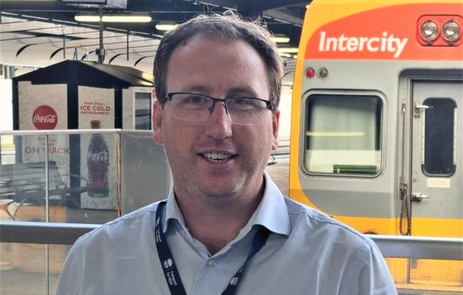 Man wearing Transport for NSW lanyard standing behind a barrier  in front of a train in a station.